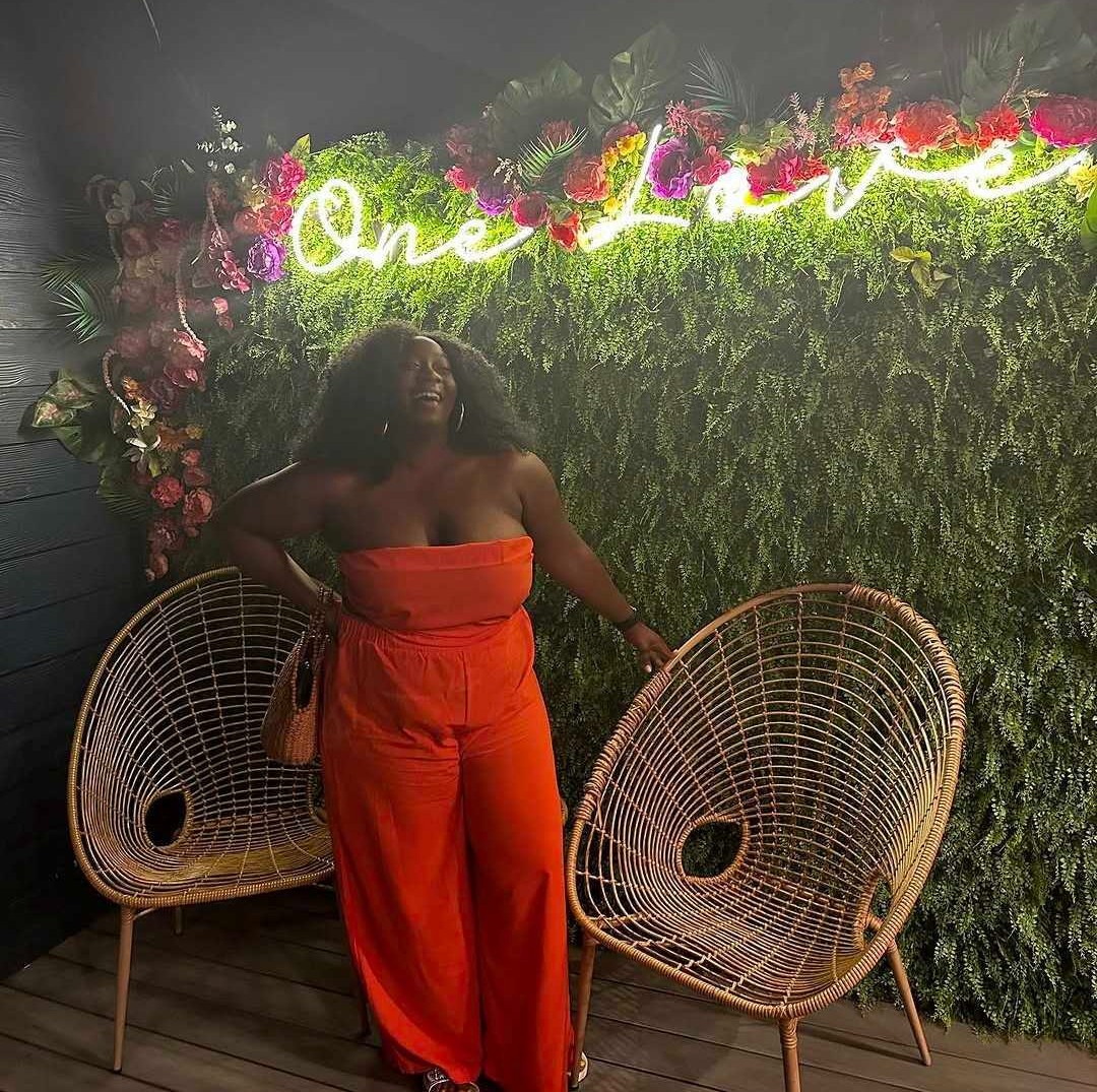 Samantha Turner smiling in a bold orange jumpsuit in front of a 'One Love' neon and a flower wall